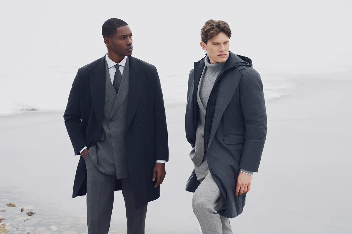 Two male models standing on a snowy beach wearing dark overcoats and gray suits.