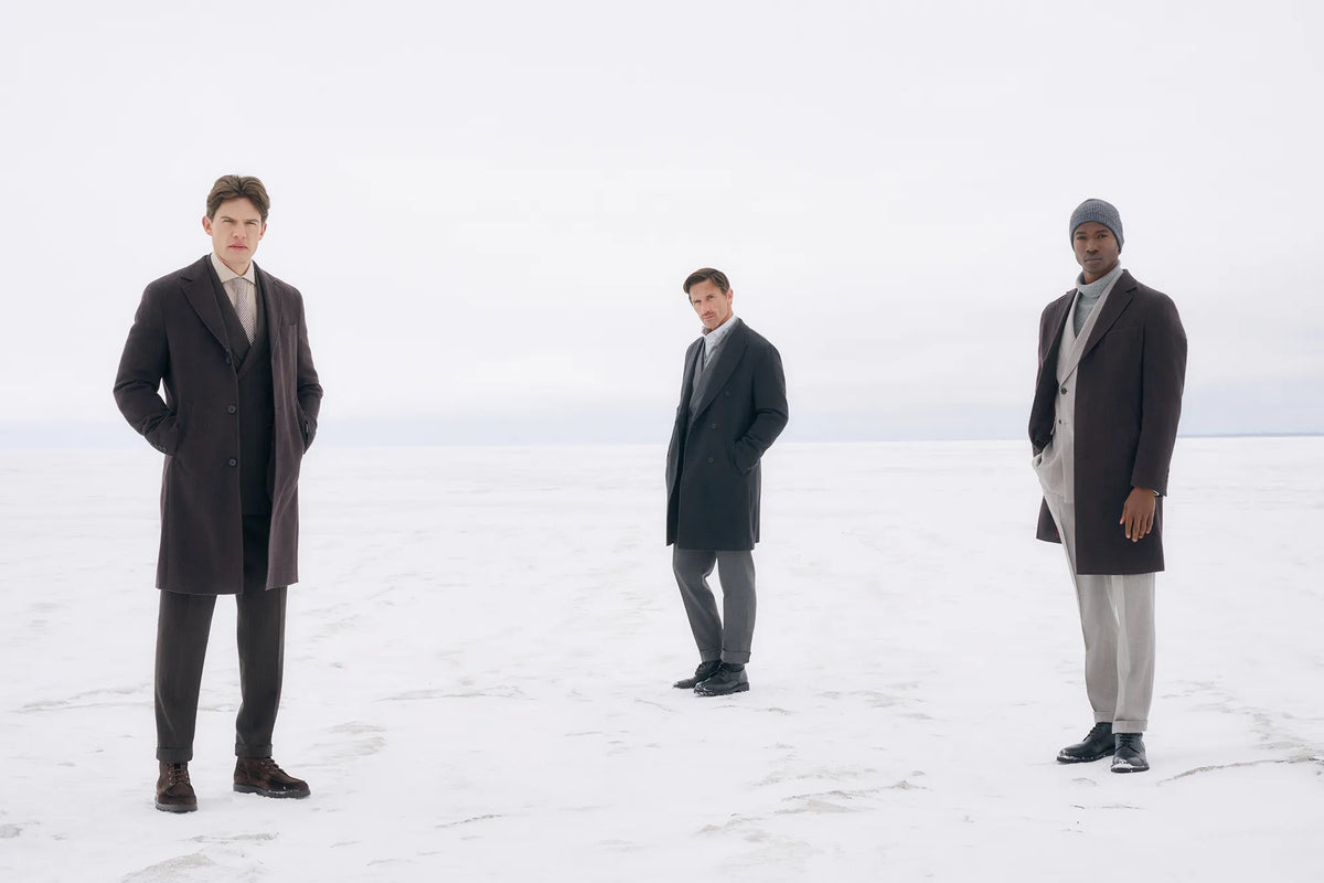 Three men standing on a snowy, overcast landscape wearing long dark overcoats and winter clothing. The image has a very bright, high-key, washed-out look with high exposure.