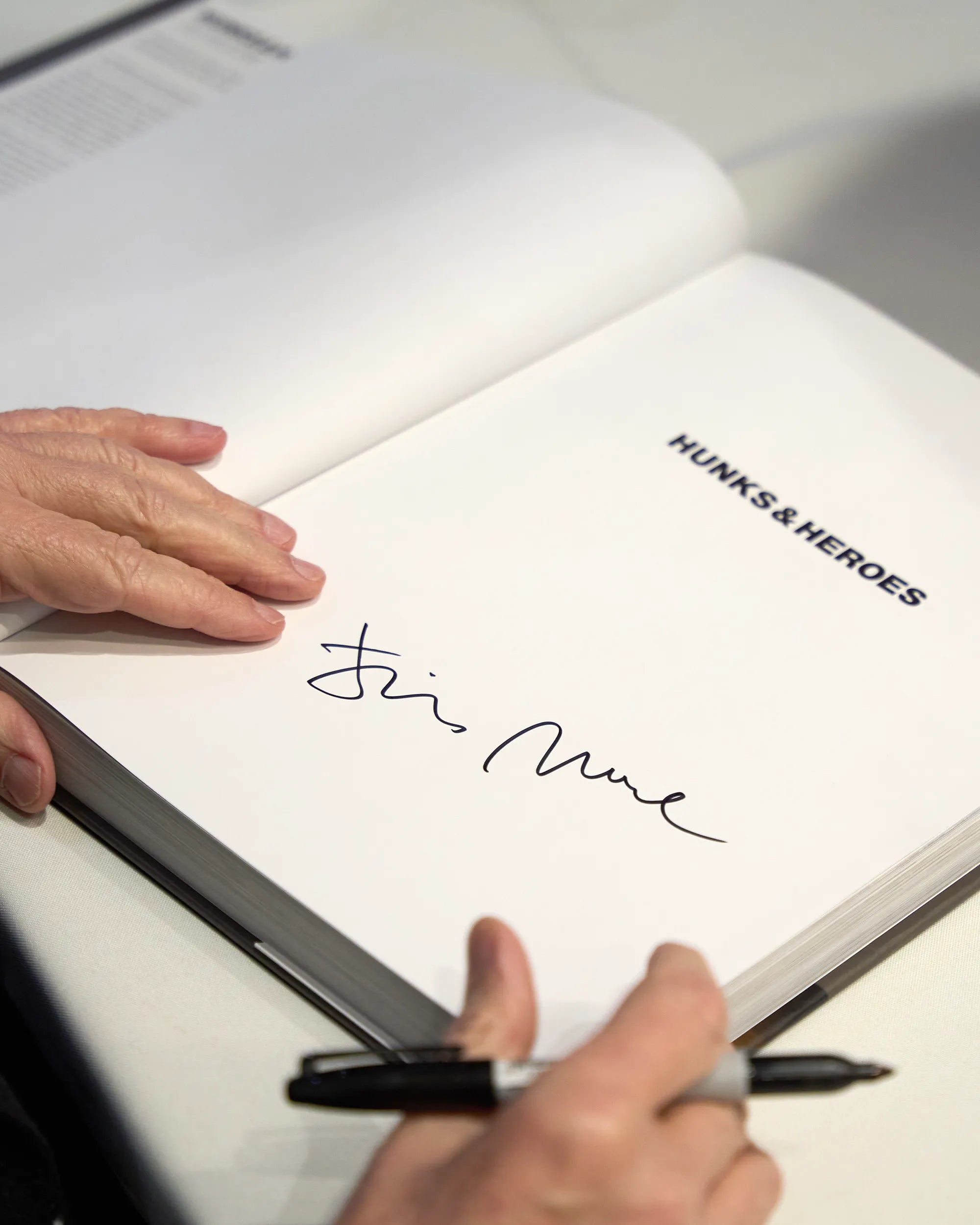 A closeup of Jim Moore's hands signing his book called 'Hunks & Heroes' on a white table.