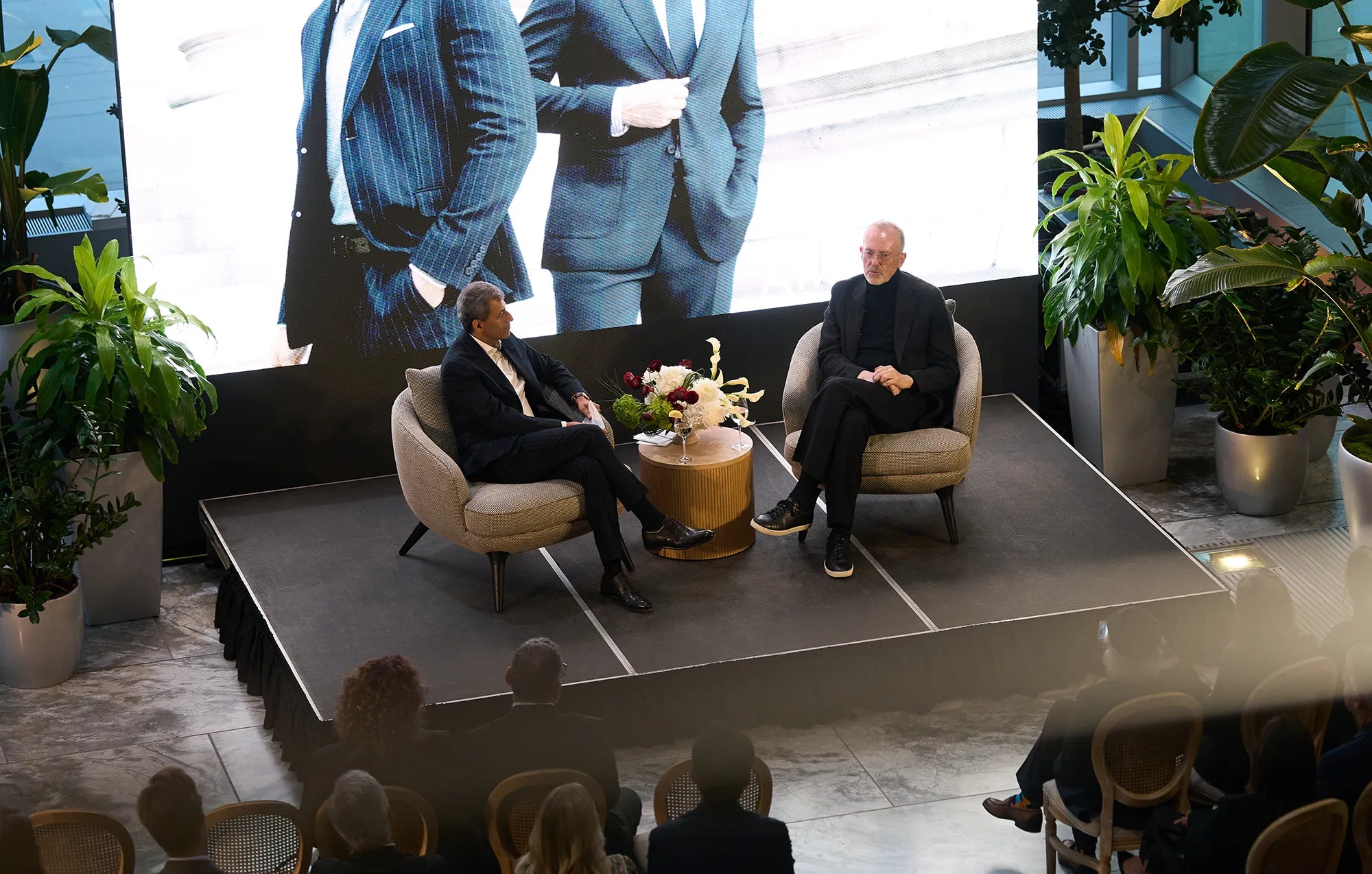 Two men in suits sitting on a stage with a large screen behind them showing models posing in a suit.