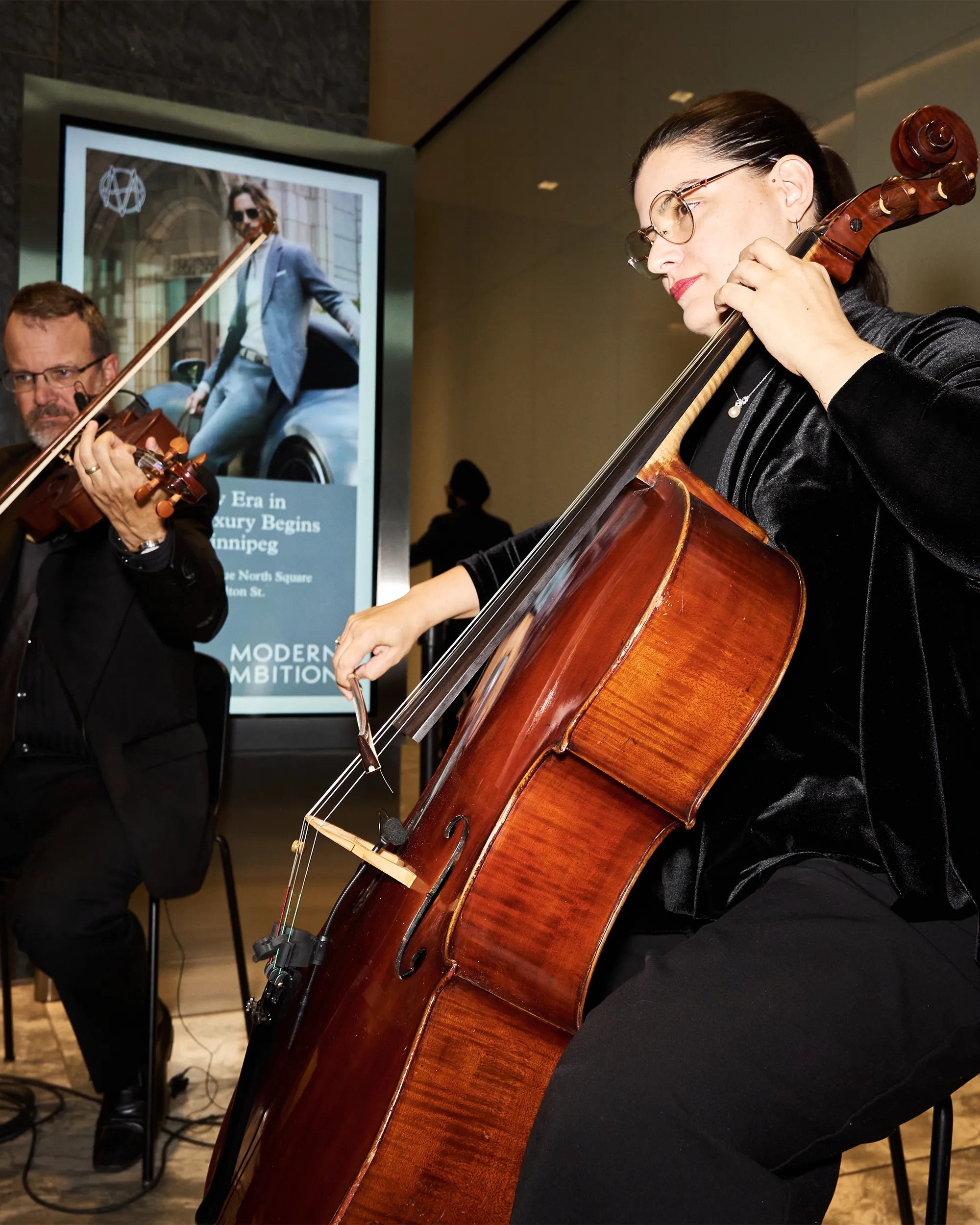 Person playing a cello with another person playing violin in the background, with a screen displaying an advertisement.