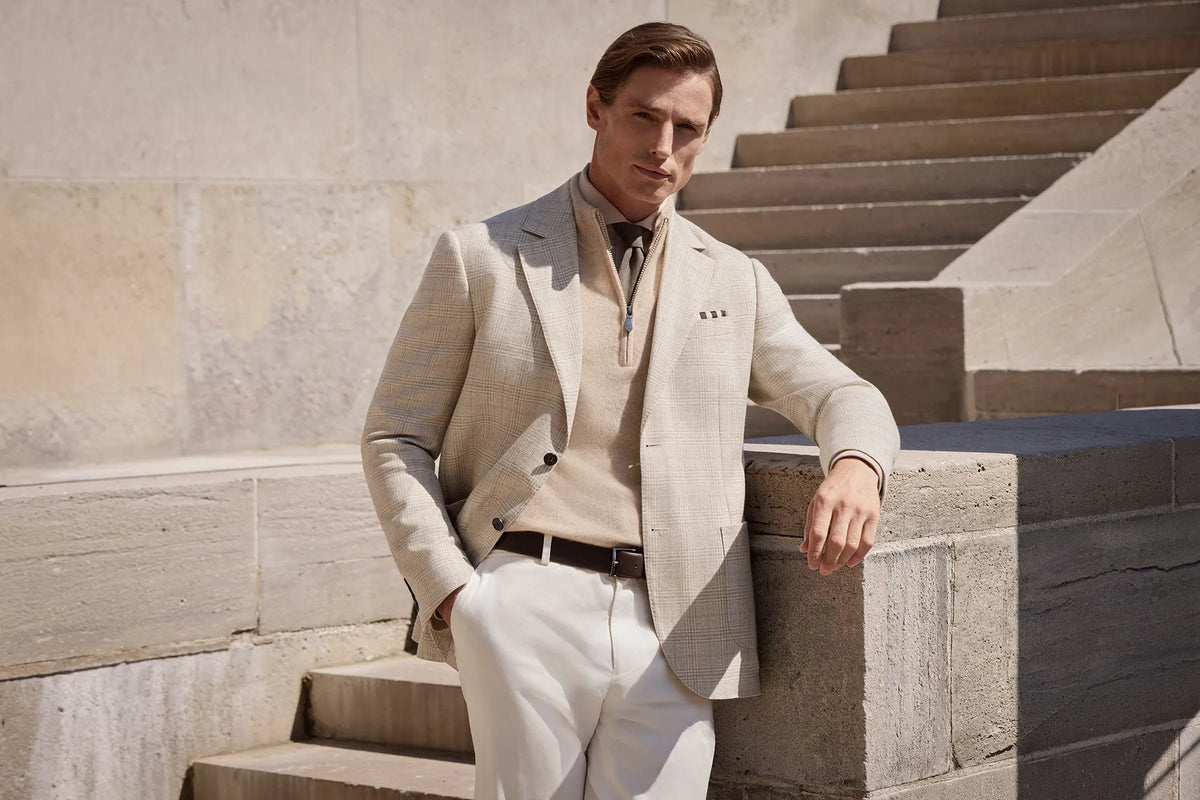 A man wearing a light beige textured blazer, cream knit sweater, white trousers, and a dark tie, leaning against a stone wall on outdoor concrete steps.